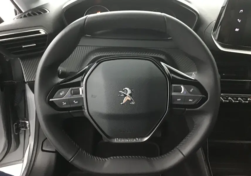 Vue rapprochée du volant cuir noir de la Peugeot 208 gris Artense, avec commandes intégrées et tableau de bord partiel visible.