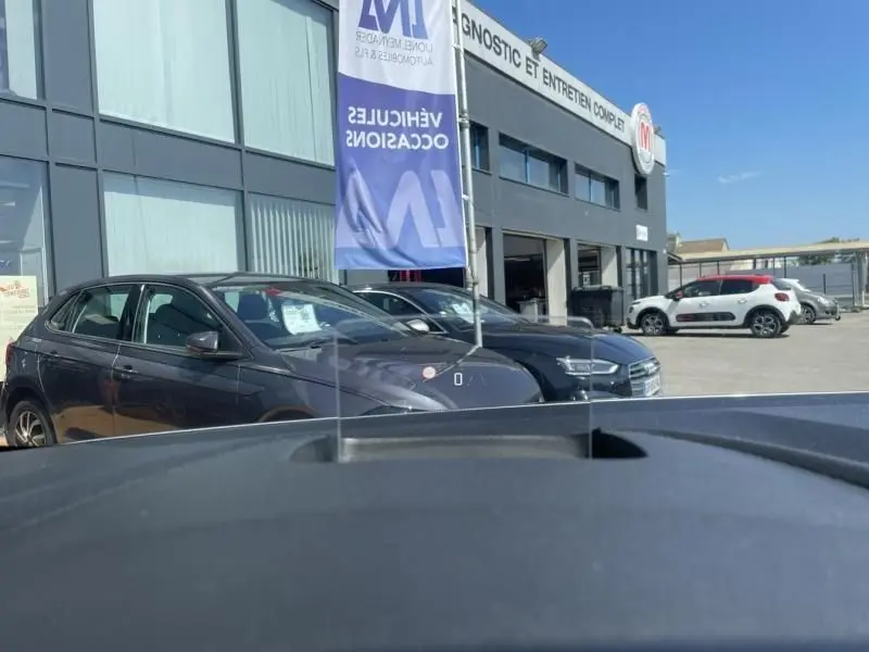 Vue 3/4 avant gauche d'un Citroën Berlingo gris métal 2018 devant un garage avec drapeau bleu visible.