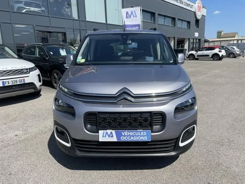 Vue de face d'un Citroën Berlingo gris métal 2018 avec calandre noire et plaque "Véhicules Occasions" devant un garage.