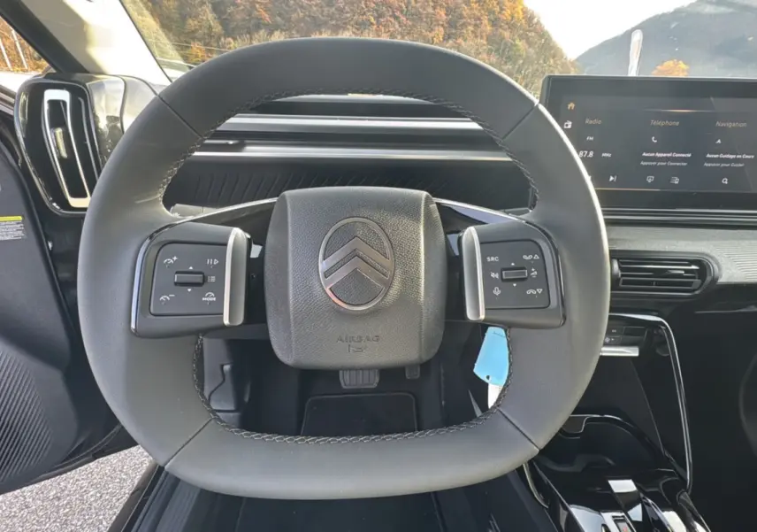 Vue rapprochée du volant cuir multifonctions Citroën C3 Aircross noir perla nera avec écran tactile en arrière-plan.