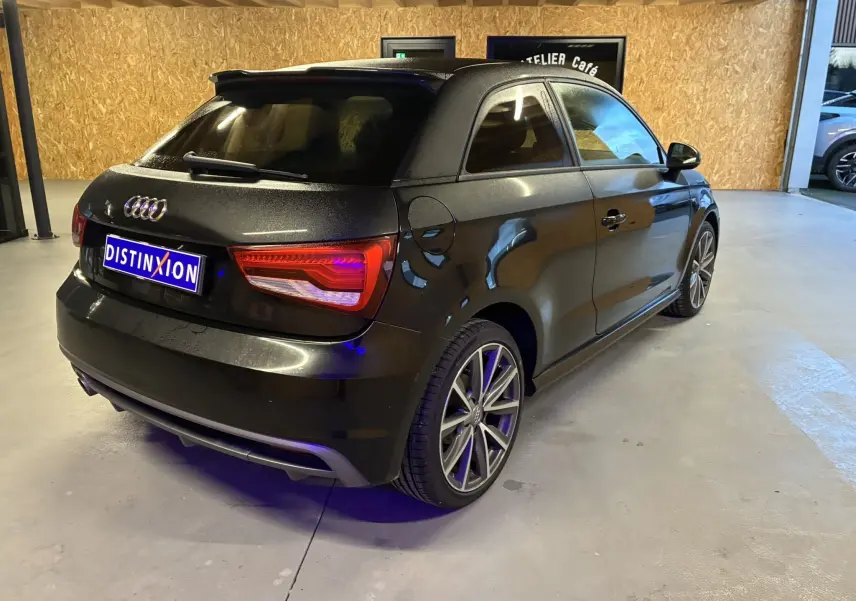 Audi A1 noire vue en 3/4 arrière droit dans un garage, avec jantes alliage et feux arrière allumés.