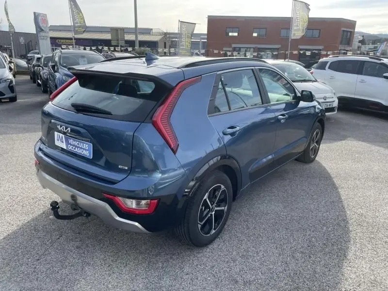 Vue 3/4 arrière droite d'un KIA NIRO 2024 bleu métal avec feux arrière distinctifs en forme de boomerang et attelage visible.
