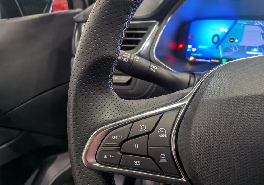 Gros plan sur le volant cuir noir perforé côté gauche du Renault SYMBIOZ E-TECH avec tableau de bord digital bleu en arrière-plan.