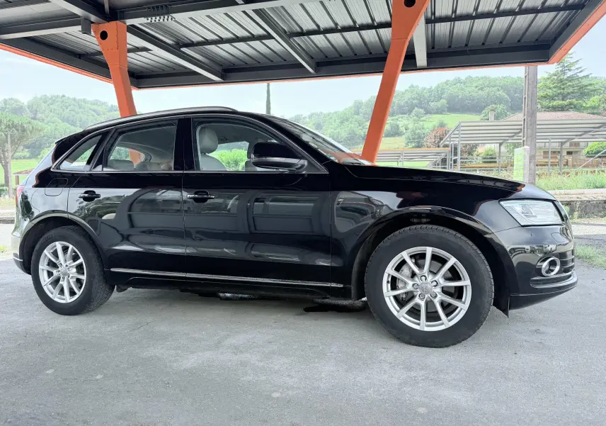 Audi Q5 noir vue de profil côté gauche, stationné sous un abri avec jantes argentées et intérieur clair visible.