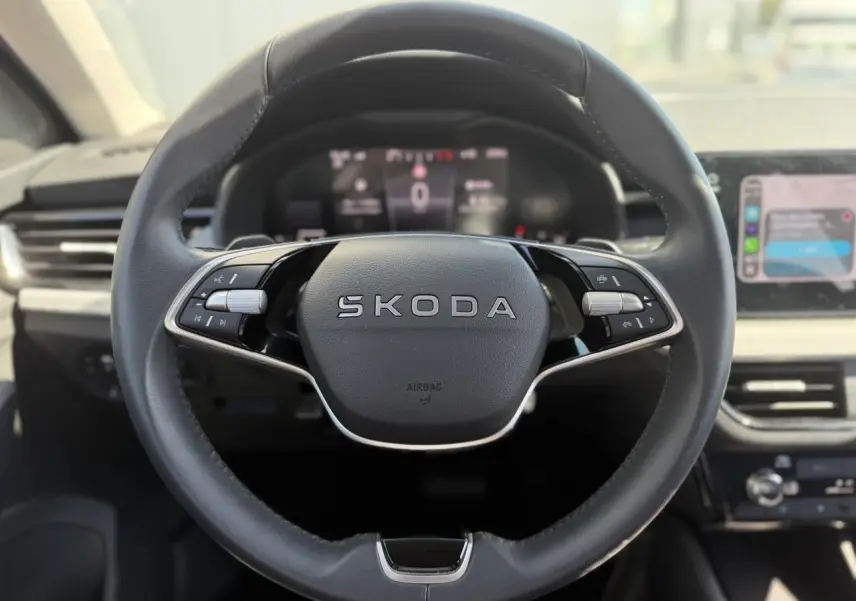 Volant cuir multifonctions SKODA en vue frontale avec tableau de bord numérique et écran tactile à droite flou.