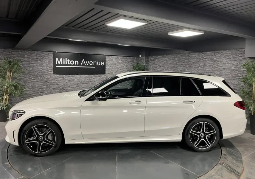Profil côté gauche d'un break Mercedes Classe C blanc avec jantes AMG noires et toit ouvrant visible en intérieur showroom.