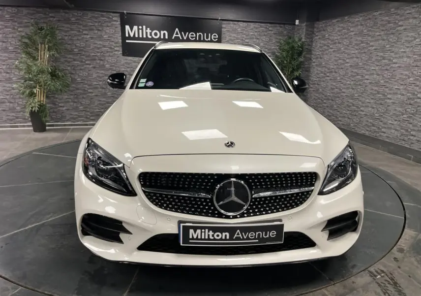 Vue frontale d'un break Mercedes Classe C blanc avec calandre AMG et logo étoile au centre, en intérieur showroom.