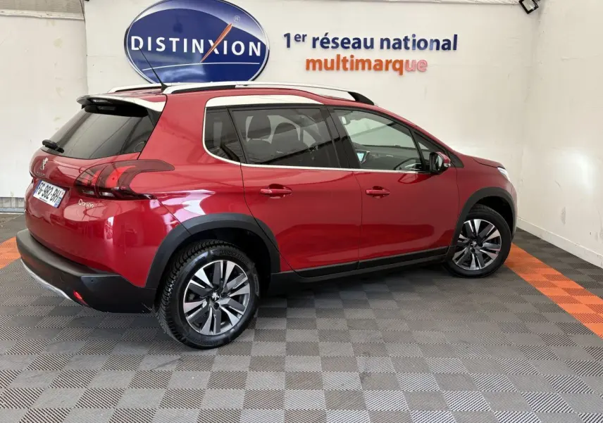 Vue 3/4 arrière droite d'un Peugeot 2008 rouge 2019 avec jantes alliage et barres de toit chromées dans un showroom.