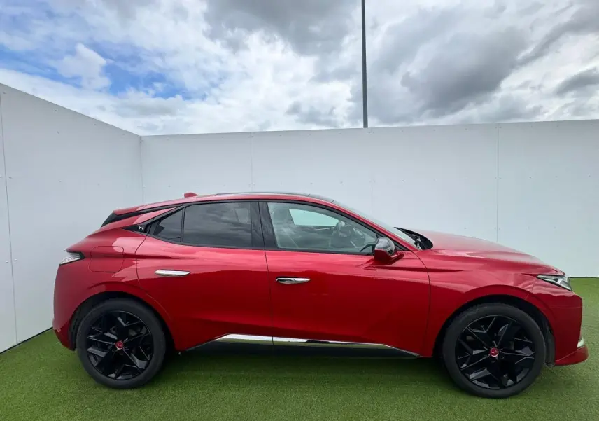 Profil côté gauche d'une DS4 rouge foncé avec jantes noires et toit panoramique sous un ciel nuageux.