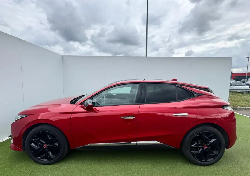 Vue latérale gauche d'une DS4 rouge foncé avec jantes noires et détails chromés sous un ciel nuageux.