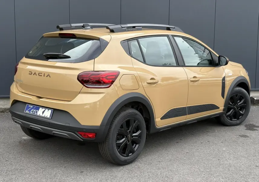 Vue 3/4 arrière droite d'une Dacia Sandero Stepway beige Safari avec jantes noires et barres de toit grises.