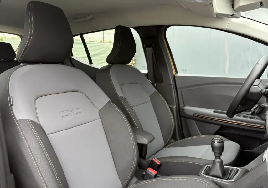 Vue intérieure côté droit des sièges avant gris clair et foncé avec accoudoir, volant et levier de vitesses manuelle visibles.