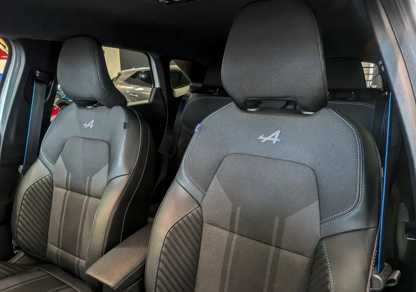 Gros plan sur les sièges avant noirs avec surpiqûres bleues et logo Alpine dans l’habitacle du Renault Symbioz blanc nacré.