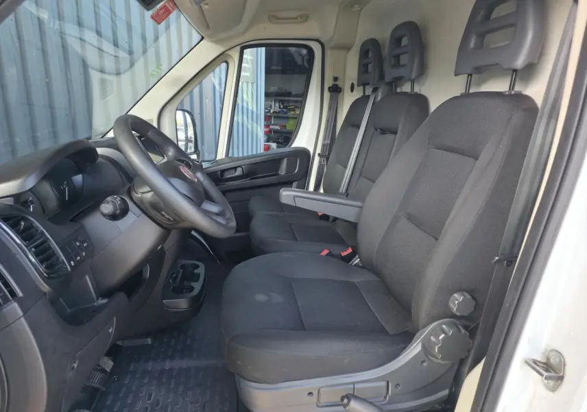 Vue intérieure côté conducteur du Fiat Ducato Fourgon blanc 2023, montrant les sièges noirs et le volant avec tableau de bord.