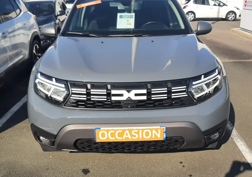 Vue avant d'un Dacia Duster gris 2024 avec calandre noire et plaque "OCCASION" visible sur un parking extérieur.