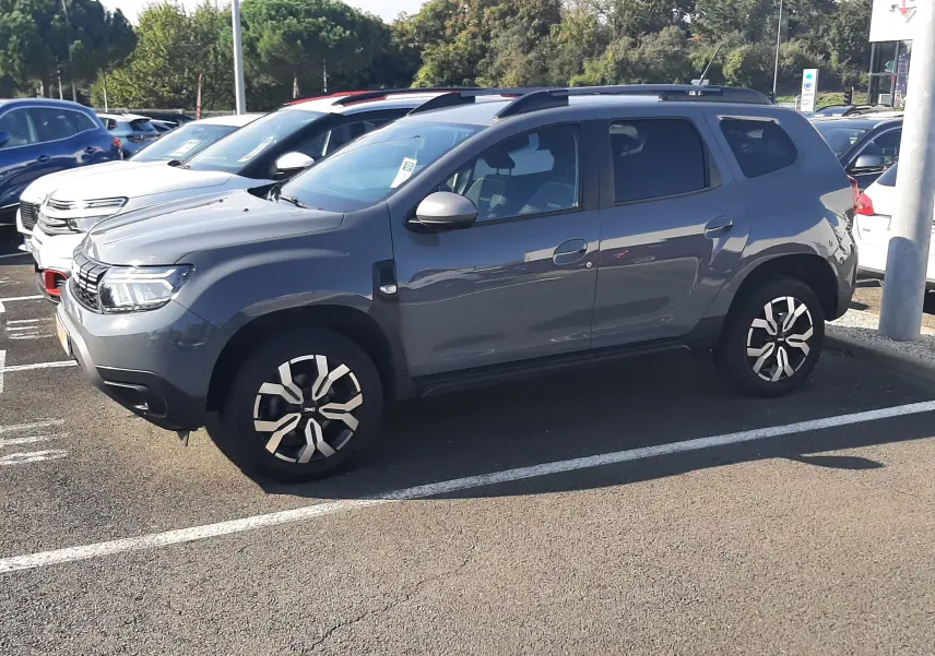 SUV Dacia Duster gris vu de profil côté gauche, avec jantes bi-ton et barres de toit noires.