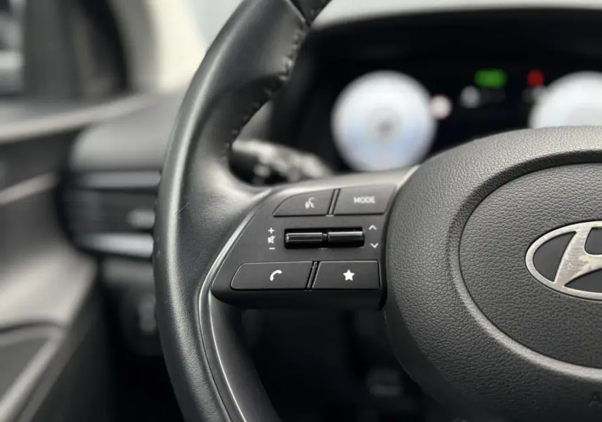 Gros plan sur le volant cuir noir de la Hyundai i20 2025 avec commandes intégrées, tableau de bord numérique flou en arrière-plan.