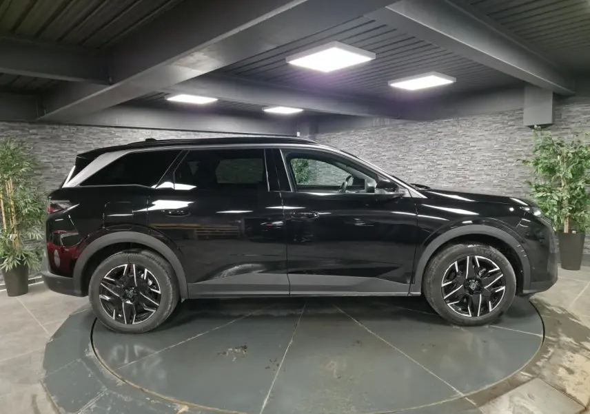 Vue de profil côté gauche d’un Peugeot 5008 noir 2025 avec jantes alliage noires et barres de toit noires brillantes dans un showroom.
