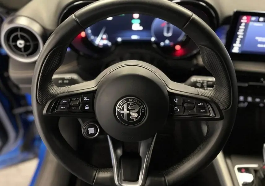 Volant sport en cuir noir avec logo Alfa Romeo au centre, tableau de bord digital et partie de la portière bleu visible.