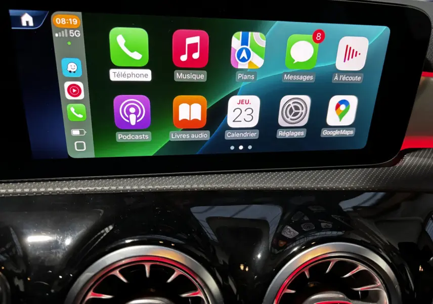 Écran tactile central affichant des applications, avec sorties d'air rondes noires et touches rouges en reflet sur tableau de bord noir brillant.