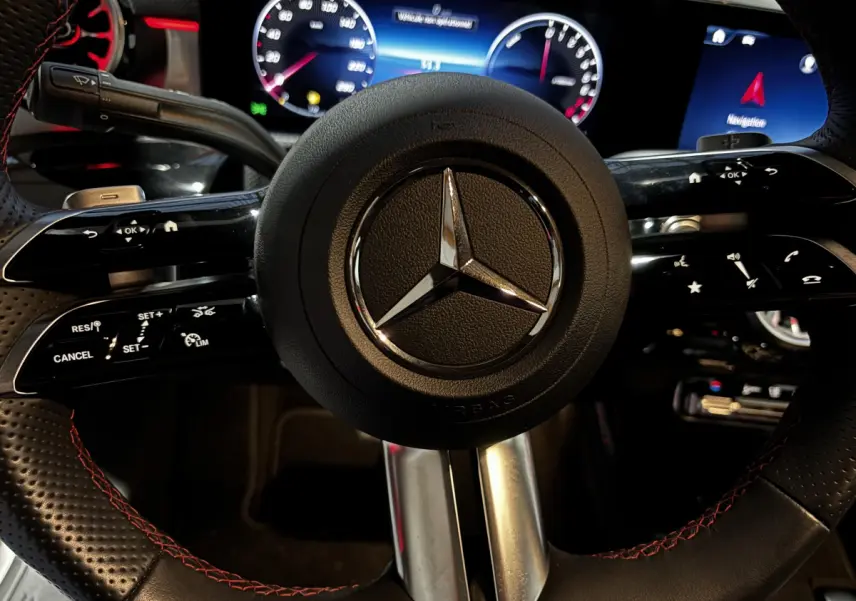 Gros plan sur le volant cuir noir avec surpiqûres rouges de la Mercedes CLA 250 e AMG Line, tableau de bord digital en arrière-plan.