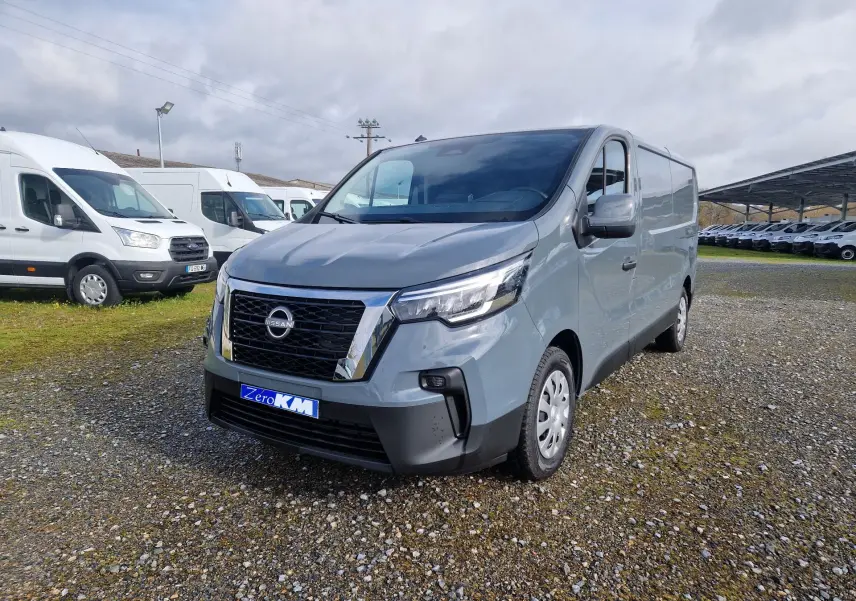 Nissan Primastar gris urbain vue 3/4 avant droit sur parking avec plusieurs fourgons blancs en arrière-plan.