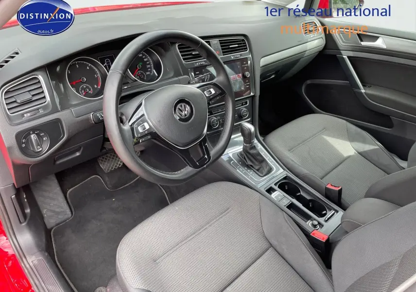 Vue intérieure côté conducteur d'une Volkswagen Golf rouge, montrant le volant, le tableau de bord et les sièges en tissu gris.