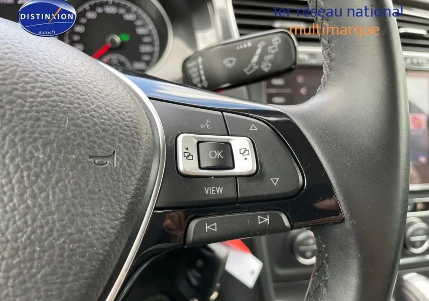 Gros plan sur les commandes au volant côté gauche d’une Volkswagen Golf rouge, intérieur cuir noir visible en arrière-plan.