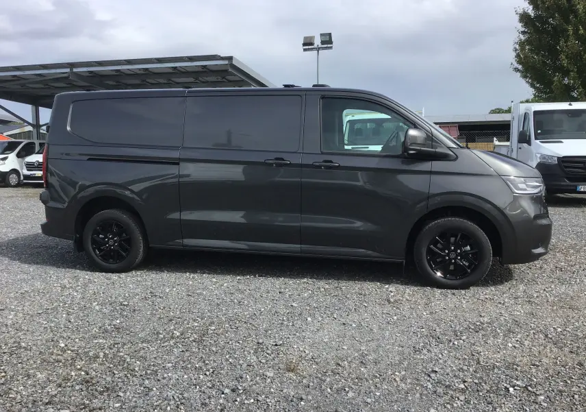 Profil côté gauche d'un Volkswagen Transporter T7 gris anthracite avec jantes noires, stationné sur un sol gravillonné.
