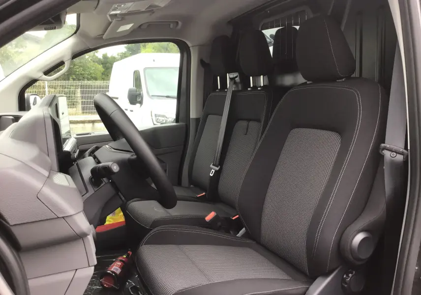 Vue intérieure côté conducteur du Volkswagen Transporter T7 2025, sièges tissu gris anthracite avec appuie-têtes et ceintures visibles.