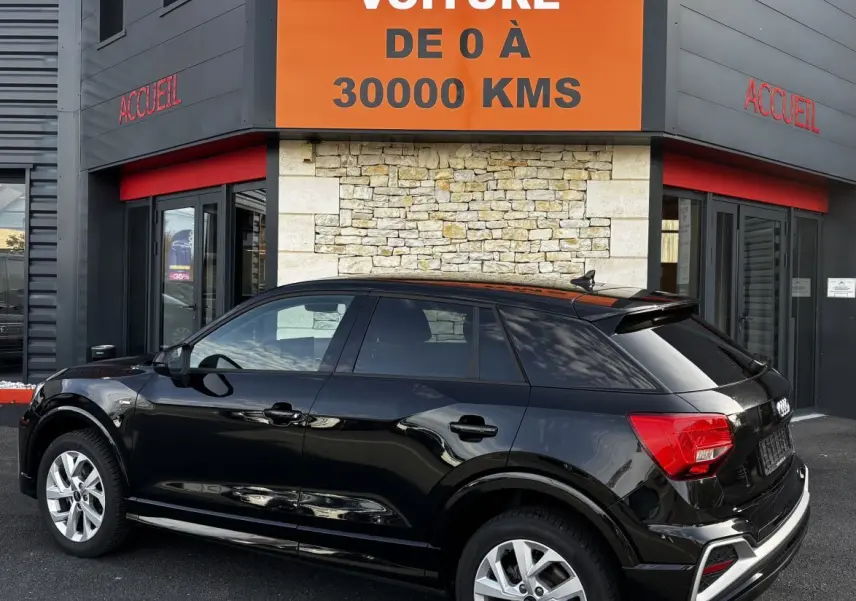 Audi Q2 35 TFSI S Line noir vue 3/4 arrière droit, stationnée devant un bâtiment avec panneau orange.