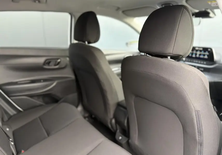 Vue intérieure arrière côté gauche de la Hyundai i20 2025, sièges tissu gris foncé et tableau de bord avec écran tactile.