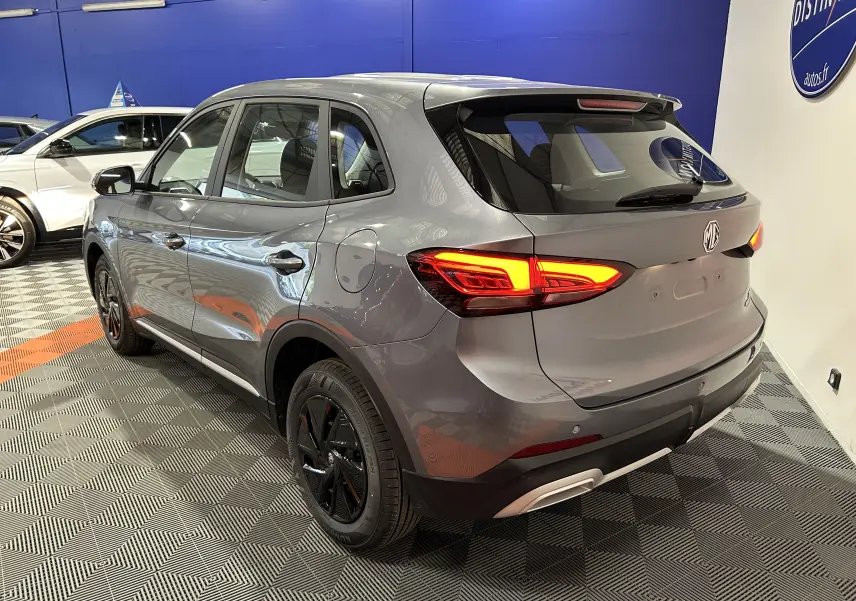 Vue 3/4 arrière droite du MG ZS Hybrid+ 2025 en gris Hampstead avec feux arrière allumés dans un showroom.