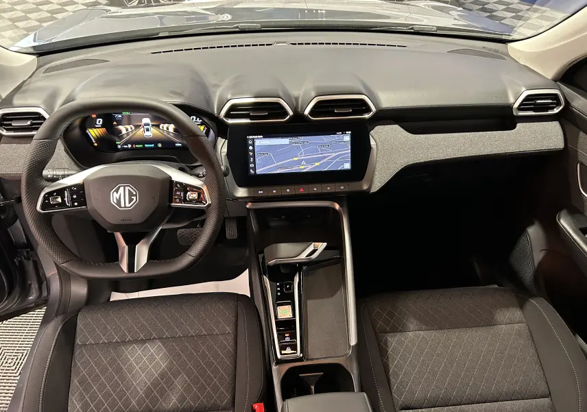 Vue intérieure frontale du tableau de bord et volant du MG ZS Hybrid+ 2025 en tissu gris avec écran tactile GPS allumé.