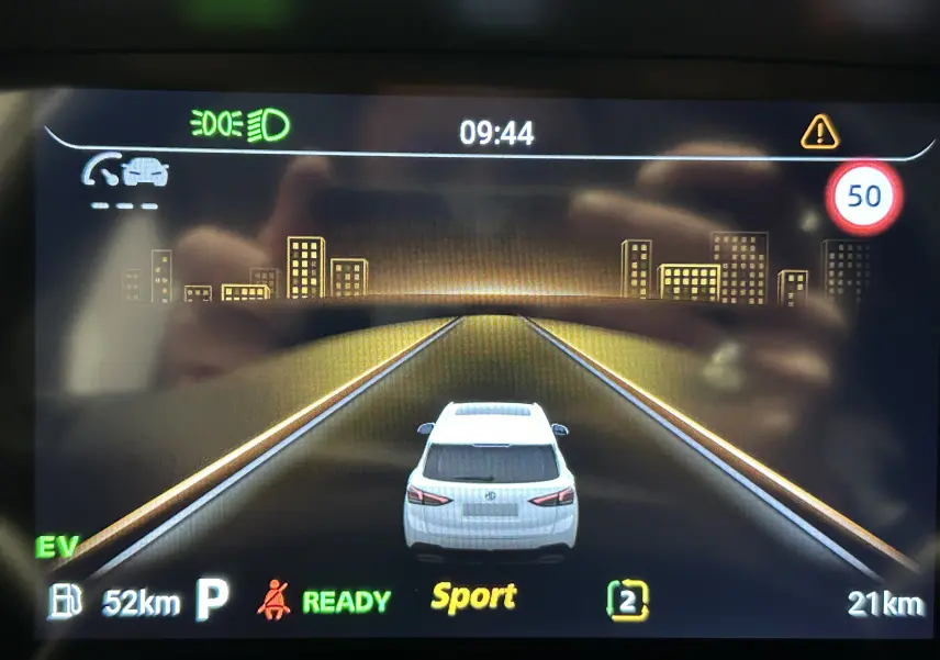 Affichage numérique montrant la voiture blanche MG ZS Hybrid+ vue de l'arrière sur une route virtuelle avec limite à 50 km/h.