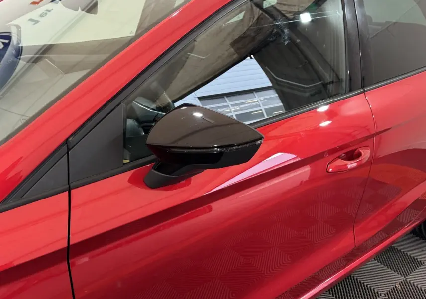 Vue rapprochée du côté gauche d’une SEAT Ibiza 1.0 TSI 110 FR rouge, mettant en valeur le rétroviseur noir et la poignée de porte.
