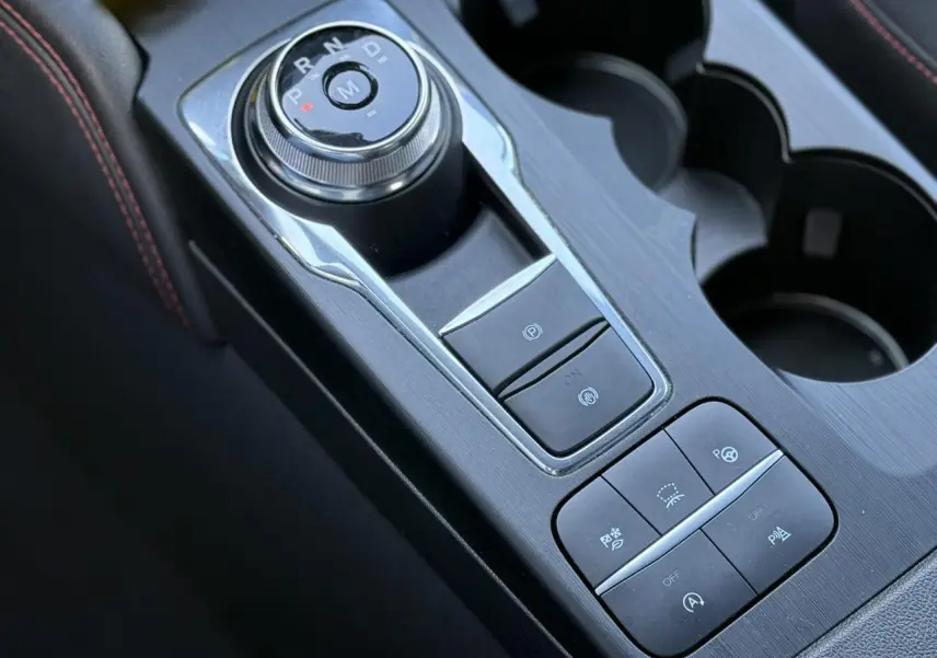 Gros plan sur la console centrale noire du Ford Kuga 2024 avec sélecteur rotatif de vitesse et boutons d’assistance au stationnement.