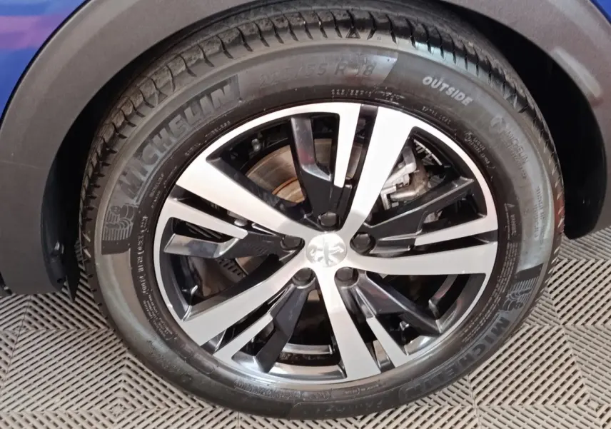 Gros plan sur la roue avant droite d’un Peugeot 3008 bleu, jante alliage bicolore et pneu Michelin visible.