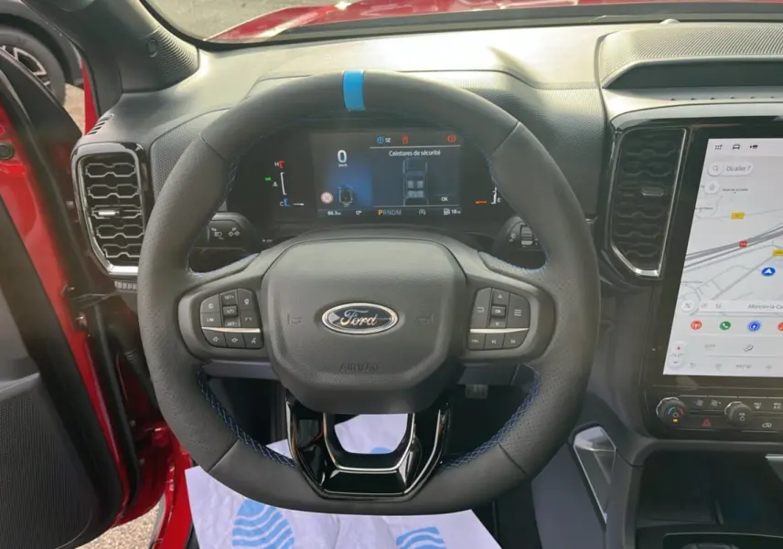 Vue rapprochée du volant cuir noir avec surpiqûres bleues du Ford Ranger 3.0 V6 Lucid Red, tableau de bord numérique allumé.