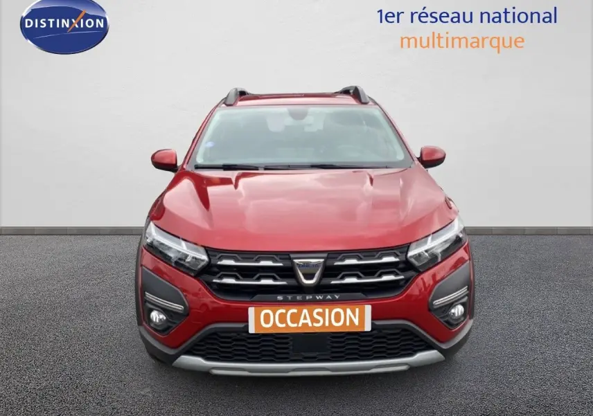 Vue frontale d'une Dacia Sandero Stepway rouge fusion métal avec calandre noire et badge central, en arrière-plan un mur blanc.