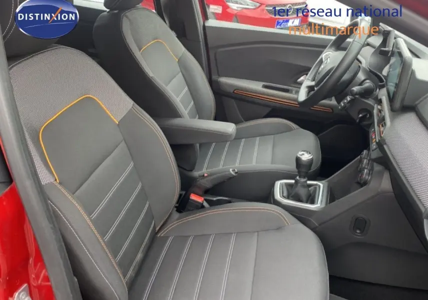 Vue intérieure côté conducteur de la Dacia Sandero rouge fusion, montrant les sièges noirs avec surpiqûres orange et la boîte manuelle.