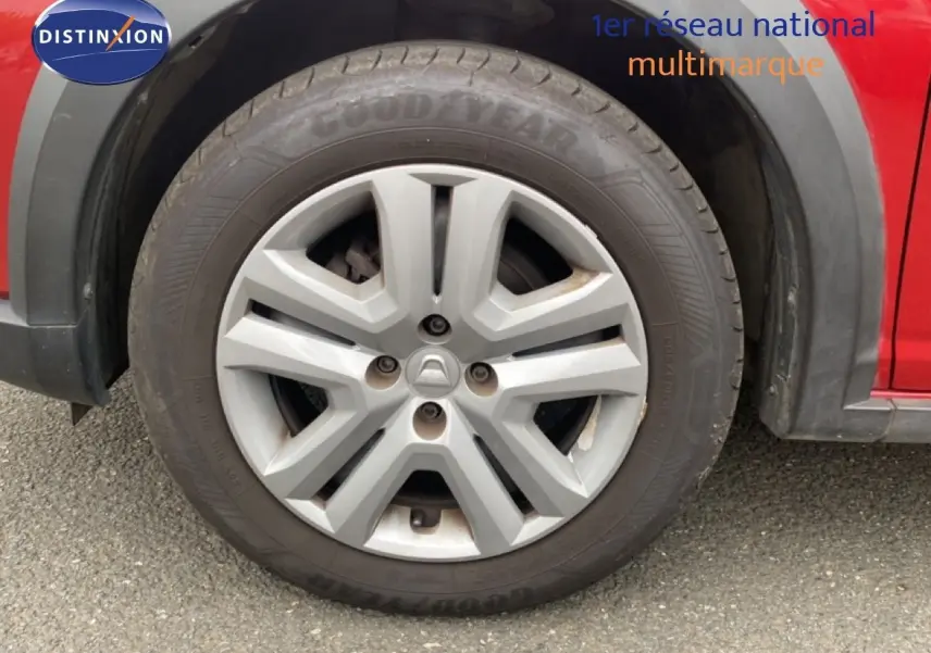 Gros plan sur la roue avant gauche d'une Dacia Sandero rouge fusion métal, jante alliage grise à six branches.