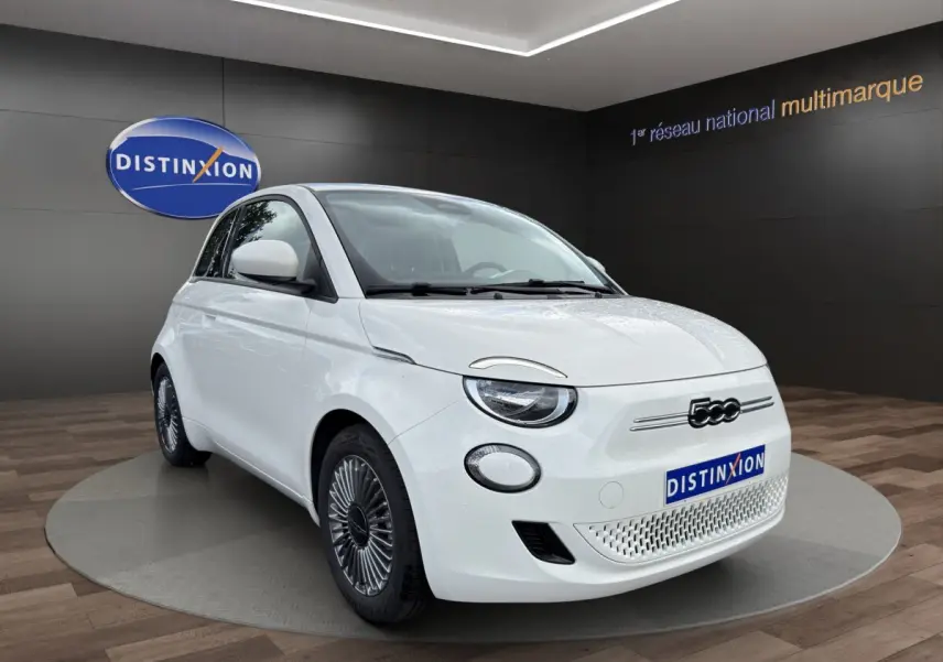 Vue 3/4 avant d'une FIAT 500 électrique blanche avec jantes alu, sur fond intérieur showroom Distinxion.