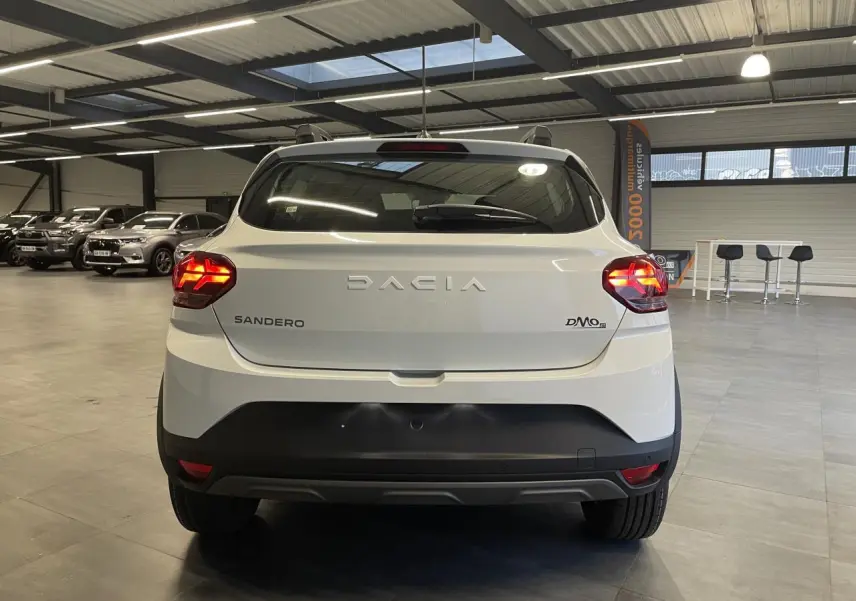 Vue arrière d'une Dacia Sandero TCe 90 Stepway blanc glacier avec feux allumés et bouclier noir en intérieur showroom.