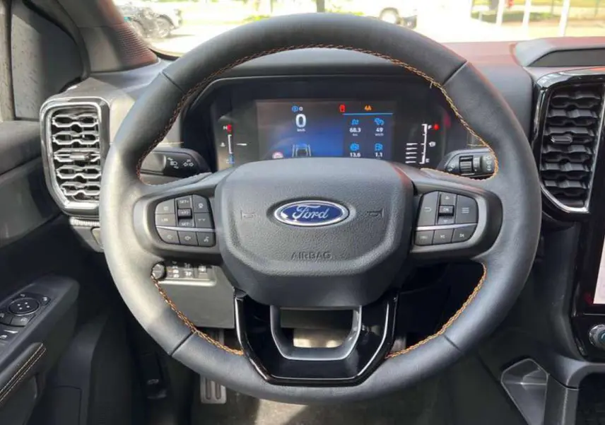 Vue rapprochée du volant cuir noir avec surpiqûres orange du Ford Ranger 2025, tableau de bord numérique en arrière-plan.