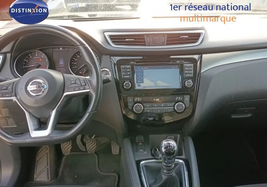 Vue intérieure centrée sur le tableau de bord et la console centrale du Nissan Qashqai blanc, avec volant et levier de vitesse manuel visibles.