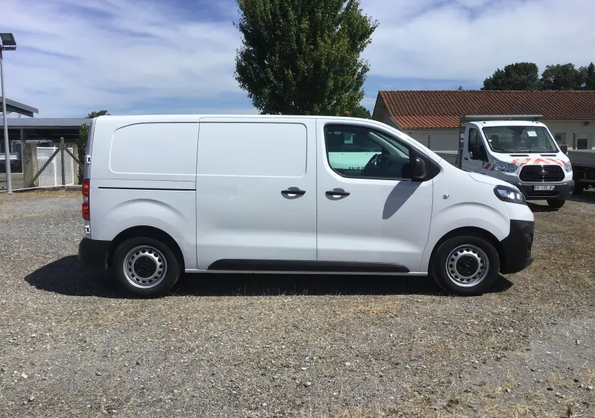 Profil côté droit d'un utilitaire blanc Opel Vivaro 2023 avec porte latérale coulissante, stationné sur sol gravillonné.