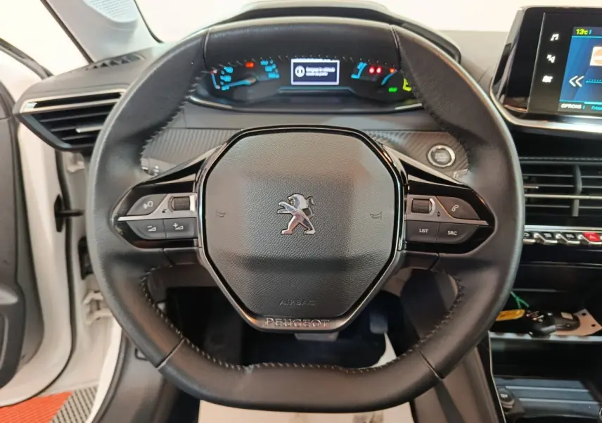 Vue rapprochée du volant cuir multifonctions d’une Peugeot 208 électrique blanche, avec tableau de bord et écran tactile en arrière-plan.