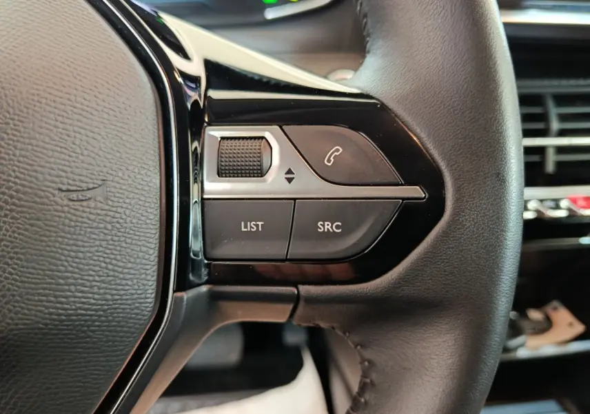 Gros plan sur les commandes gauche du volant cuir noir du Peugeot 208 électrique blanc, avec boutons multifonctions.
