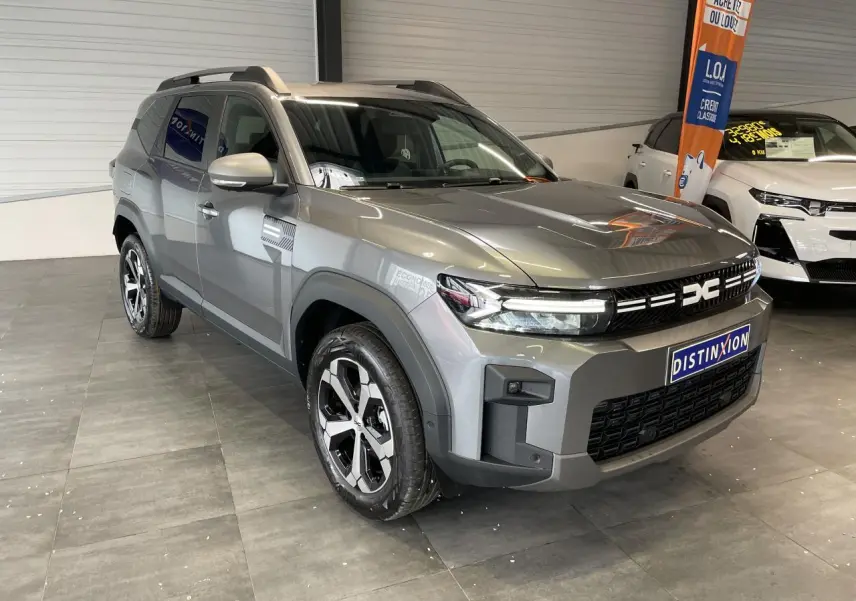 SUV Dacia Bigster gris schiste en 3/4 avant droit, avec jantes alliage et barres de toit visibles en intérieur showroom.